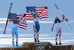 Photographie de trois skieuses sur un podium, deux d'entre elles brandissant le drapeau des États-Unis, l'autre portant ses skis.