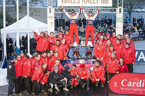 Sébastien Loeb et Daniel Elena debout sur le toit de leur Citroën C4 WRC, levant les bras au ciel et entourés de l'équipe de Citroën Racing.