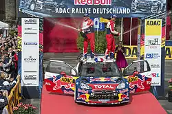 Sébastien Loeb et Daniel Elena sabrant le champagne debout sur le toit de leur Citroën DS3 WRC, portières ouvertes et entourée de spectateurs.
