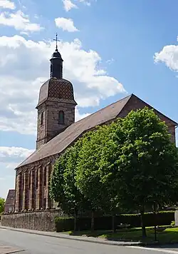 Vue arrière d'une grande église en grès rose surmontée d'un clocher comtois et d'un carillon.