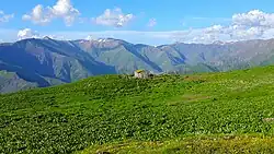 Une cabane avec petit jardin dans des alpages ; derrière, une chaine de montagnes.
