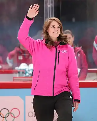 Photographie d’une femme, saluant de la main droite, portant une veste rose avec les anneaux olympiques.