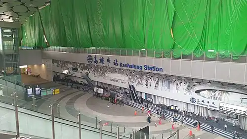 Chantier d'une vaste halle dont le mur est couvert d'une bâche verte.