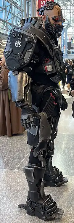 Homme de plein-pied portant une armure noire et grise, et un casque rouge et noir.
