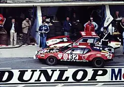 Photo d’époque d’une voiture de course rouge dans la ligne des stands du Mans
