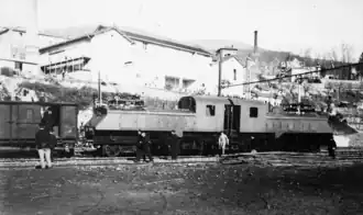Photographie en noir et blanc de la locomotive vue de profil devant les grandes constructions des parfumeries de Grasse.