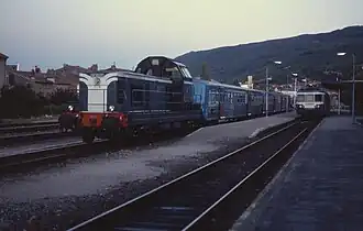 Photo couleur d'une locomotive diesel mono cabine bleu et blanche attelée à une rame bleu et inox à quai à gauche. À droite, un autorail bleu et blanc est de face.