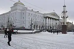 Bâtiment majestueux à colonnes blanches avec des murs bleu ciel et un dôme doré entouré de rues enneigées. À l'avant-plan, des piétons traversent une place enneigée tandis qu'une limousine blanche allongée est garée près du trottoir.