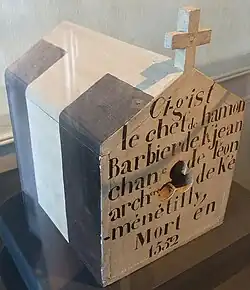 Boîte à crâne en forme de petite maison surmontée d'une croix avec une inscription dessus.