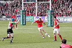 Joueur au centre, de face, en train de réaliser un coup de pied de dégagements dans son en-but, sous le regard de partenaires. Un adversaire, les bras levés, tente de le contrer.