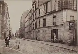 photographie sépia de la rue ; au premier plan se trouve une enseigne "crèmerie de la butte Montmartre" devant laquelle une femme est assise. Plusieurs enfants sont dehors, et des adultes se tiennent sur le pas des portes