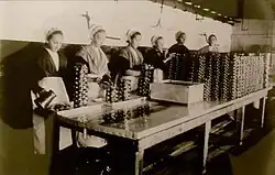 Ouvrières d'une conserverie de Douarnenez debout face à une table de travail où sont posées les boîtes dans lesquelles elles versent de l'huile, début du XXe siècle