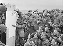 Photo en noir et blanc d'un homme en uniforme parlant à d'autres hommes, assis ou debout tout autour de lui, qui écoutent attentivement le premier en regardant la carte qu'il montre.