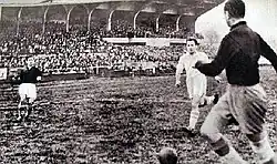 Joueur en noir avec le ballon proche de lui, avec un adversaire à quelques mètres, et un coéquipier plus loin.