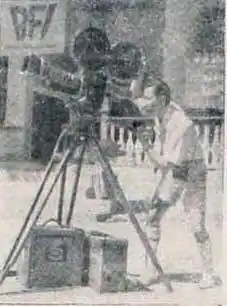 Vue d'un tournage cinématographique ancien. Un homme d'âge moyen, de type asiatique, utilise une caméra antique sur trépied, vêtu d'un habit léger des années 1930-1950. L'arrière-plan flou montre une structure en pierre. L'image en noir et blanc, avec un éclairage neutre, met l'accent sur l'homme et son équipement, créant une ambiance sobre.