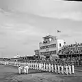 Cliché en noir et blanc. Au premier plan, probablement provenant d'un avion garé au loin à gauche, cinq personnages, dont le couple royal, passent devant un triple rang de militaires hommes au garde-à-vous. Tout le monde est en blanc et, sauf la reine, en uniforme. Au second plan à droite, bâtiment de l'aérogare, avec la tour de contrôle. Une foule est amassée au pied du bâtiment et sur ses balcons ; face à elle, un triple rang de femmes en uniforme de teinte sombre, dos tourné aux militaires hommes.
