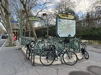 Photographie d'une bouche de métro aux rambardes de laquelle sont accrochés des vélos ; en arrière-plan, une longue perspective animée par une grille sombre aux pointes dorées.