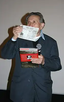 Un homme âgé montrant un billet de banque et tenant un livre dans sa main gauche.