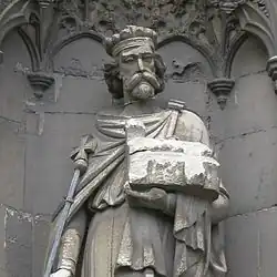 Statue en pierre grise d'un homme barbu et couronné qui tient un sceptre dans la main droite et une église en modèle réduit dans la main gauche.