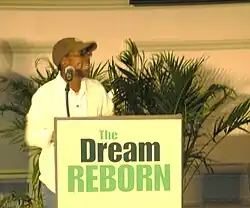 Photo d'une femme noire sur une tribune. Elle porte une chemise blanche, une casquette et des lunettes de soleil.