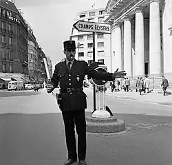Un gendarme sur une place parisienne, le sifflet à la bouche et le bâton à la main. Derrière, un panneau indiquant la direction des Champs-Élysées.