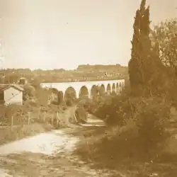 Photographie en sépia représentant le viaduc dans le fond, une petite maison à gauche, un chemin au centre et un cyprès à droite.