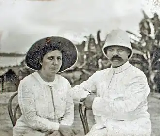Une jeune femme coiffée d'un chapeau à large bords et vêtue de blanc au côté d'un homme moustachu coiffé d'un casque colonial.