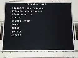 Tableau d’affichage Lectovisuel noir rainuré, en matière synthétique, encadré d’aluminium, sur lequel on a accroché des lettres (certaines manquent) pour donner le menu du 21 mars 1963 : assortiment de céréales sèches, blé entier à la vapeur, 1 œuf brouillé, 2 lait, compote de fruits, pain grillé, pain, beurre, café.