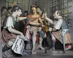 Photographie en couleurs. Groupes de statues colorées représentant Jésus entourés de soldats, dont deux accroupis de dos au premier plan.