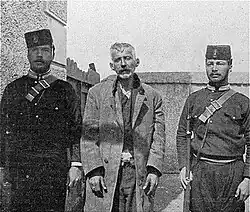 Photographie en noir et blanc montrant un homme à l'air hagard entouré de deux soldats.