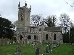 Photo d'un bâtiment allongé en pierre grise flanqué d'une grande tour carrée portant un horloge, au milieu d'une pelouse verte avec des rangées de pierres tombales