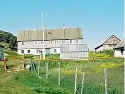 Photographie en couleurs de terrains herbus séparés d'une clôture grillagée, des bâtiments gris faits de tôles visibles au second plan.