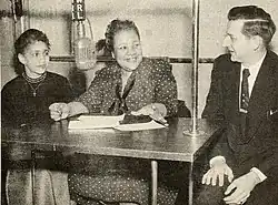 Photographie de deux femmes et d'un homme assis à une table avec un microphone suspendu au centre et des papiers sur la table.