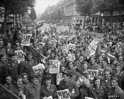 Foule de soldats alliés en liesse dans une rue ; plusieurs brandissent un journal avec le mot Piece inscrit en grand.