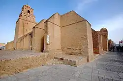 Vue extérieure de la Grande Mosquée, centrée sur l'angle nord-ouest, montrant plusieurs contreforts à toit en pente de l'enceinte, ainsi que le minaret. Les solides contreforts, qui épaulent cette dernière, sont de diverses formes et dimensions, et sont, pour la plupart, épais et saillants.