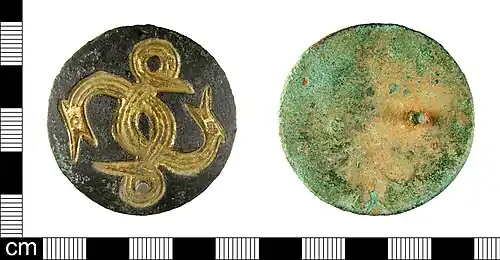 Photo des deux faces d'un disque en métal corrodé. Une face est vierge, l'autre porte un dessin gravé représentant deux serpents entrelacés dont le corps fait une boucle avec la queue en face de leur gueule grande ouverte.