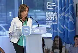 Photographie d'une femme debout derrière un pupitre, portant une veste blanche et prononçant un discours.
