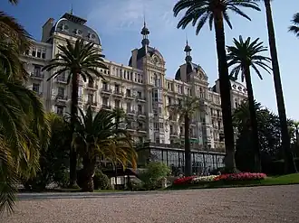 Photographie en couleur d'une grande façade claire et ouvragée sur fond de ciel bleu, avec des palmiers