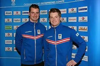 Photographie de deux hommes portant la même veste de survêtement bleue à parements rouges et blancs, posant devant un panneau publicitaire.