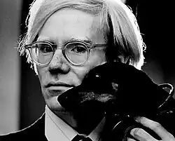 Portrait photographique en noir et blanc d'un homme blond portant des lunettes tenant dans sa main un petit chien