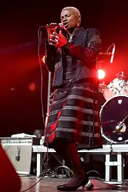 Vue en contre plongée d'une chanteuse africaine aux cheveux courts et teints en blond, vêtue d'une robe à rayures noires et grises en train de chanter sur une scène.