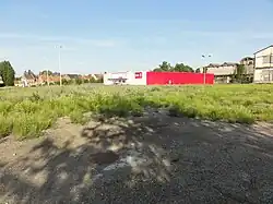 La photographie montre la tête de puits matérialisée, soit une plaque d'égout entourée d'une dalle de béton, dans son environnement, à savoir une friche. Un supermarché Dia apparaît en arrière-plan.