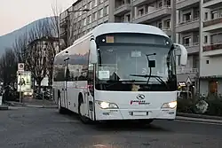 Photographie en couleurs d’un autocar blanc roulant dans une rue urbaine, devant un immeuble résidentiel.