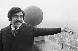 Photo en noir et blanc d'un homme brun moustachu souriant qui désigne du bras une ville qui s'étend en contrebas