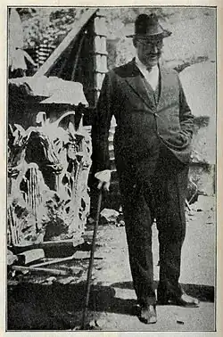 Photographie en noir et blanc et en pied d'un personnage en costume et chapeau haut-de-forme.