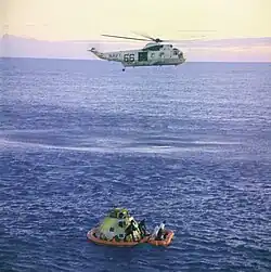 Un hélicoptère survole un vaisseau spatial et un canot pneumatique flottant sur la mer.