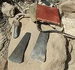 Posés sur une pierre plate, au premier plan, deux lames ou grattoirs en métal, au deuxième plan, un autre outil rattaché à une corde et une besace en cuir.