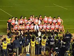 équipe de rugby posant devant des journalistes. Les joueurs sont sur deux rangs, ceux du premier rang étant assis.