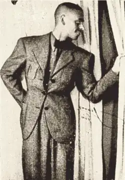 Photographie en noir et blanc d'un homme debout vêtu d'un complet. De sa main gauche il écarte un rideau et regarde ce qui se passe à travers cette ouverture. Son autre main se trouve derrière son dos.