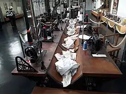 Photographie en vue rapprochée d'une rangée de postes de travail des ouvrières chemisières du XIXe&nbsp;siècle reconstitués au musée. Un unique meuble long en bois accueille deux rangées de places qui se font face, chacune pourvue d'une machine à coudre. Des séparateurs en métal arborant la marque Singer séparent les places. Au centre, une rigole creuse dans le bois accueille le linge. Chaque place dispose d'un tabouret.